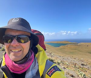 A smiling man wearing a broad brimmed hat, hot pink buff and yellow goretex jacket stands amidst a hilly green landscape with a perched lake and ocean in the background.