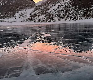 Glasslike ice, scarred by white cracks are in the foreground, with snowy and rocky mountains behind with a light pink sunset peeking between peaks.