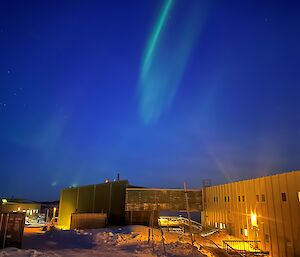 A single striking aurora is directly above a collection of green and yellow buildings, in a snowy landscape.