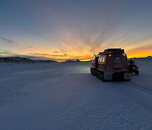 The silhouette of a Hagglungs vehicle sits in a dark snowly landscape with the last vestiges of an orange sunset being thrown above the horizon.