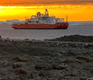 A red and white boat sits in calm waters, just off the coast with a bold golden and orange sunset in a cloudy sky