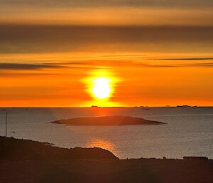 A shimmering orange sun is about to set over the horizon. In the foreground a small rocky island protrudes from a calm ocean