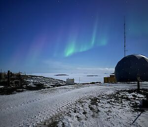 A wispy green auroro is painted in a deep blue sky above a dome and some antennas in a snowy landscape.