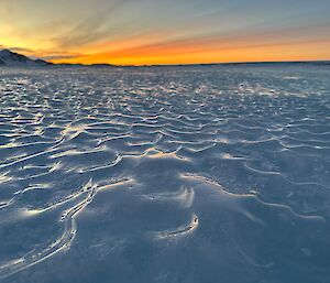 A light organce sunset appears in the distance beyond an expanse of blueish white snow with waves in it