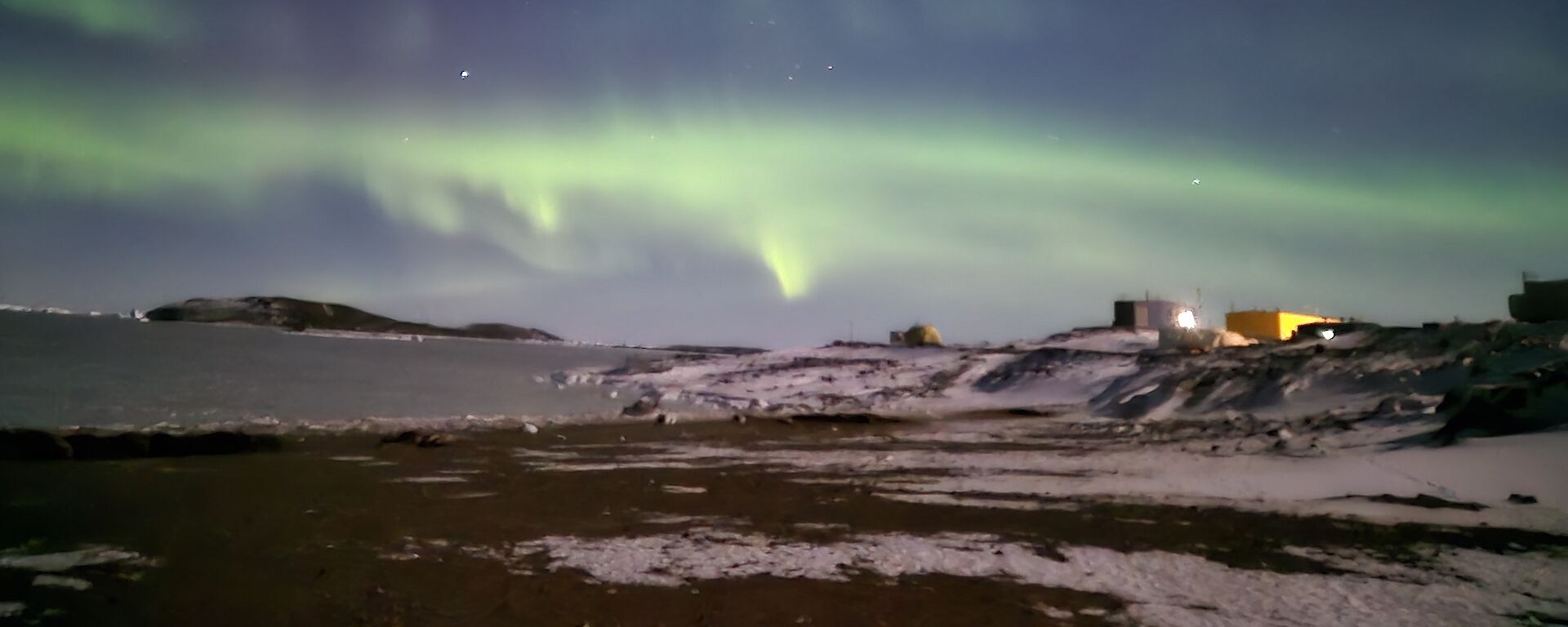 A view from a distance of some colourful buildings at Davis Station, with an icy sea with a green aurora visible in a blue sky.