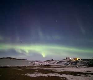 A view from a distance of some colourful buildings at Davis Station, with an icy sea with a green aurora visible in a blue sky.