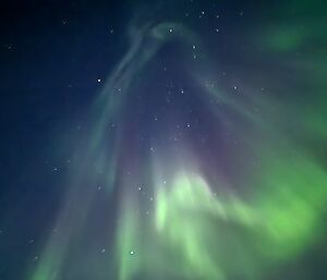 A blue sky with some stars, and a dramatic aurora that is mostly green, with some purple, covers most of the sky.