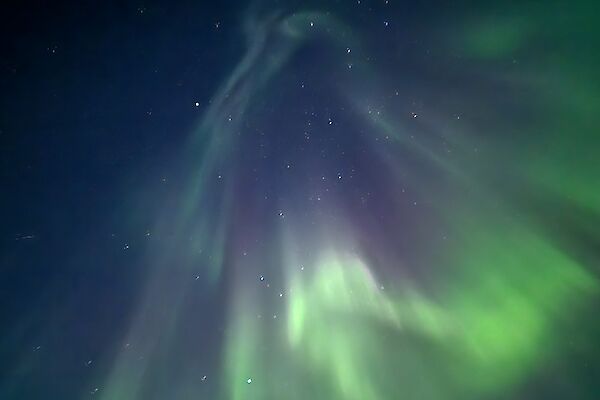 A blue sky with some stars, and a dramatic aurora that is mostly green, with some purple, covers most of the sky.