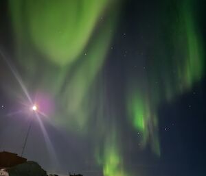 A green aurora fills most of the night sky, with a scattering of stars. There is a light on a mast in the foreground, above a snowy landscape.