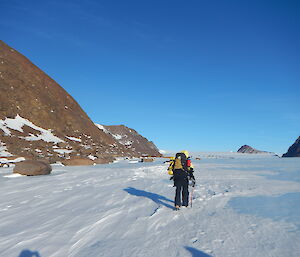 lone expeditioner walking on blue ice with rocky mountain features on left-hand side