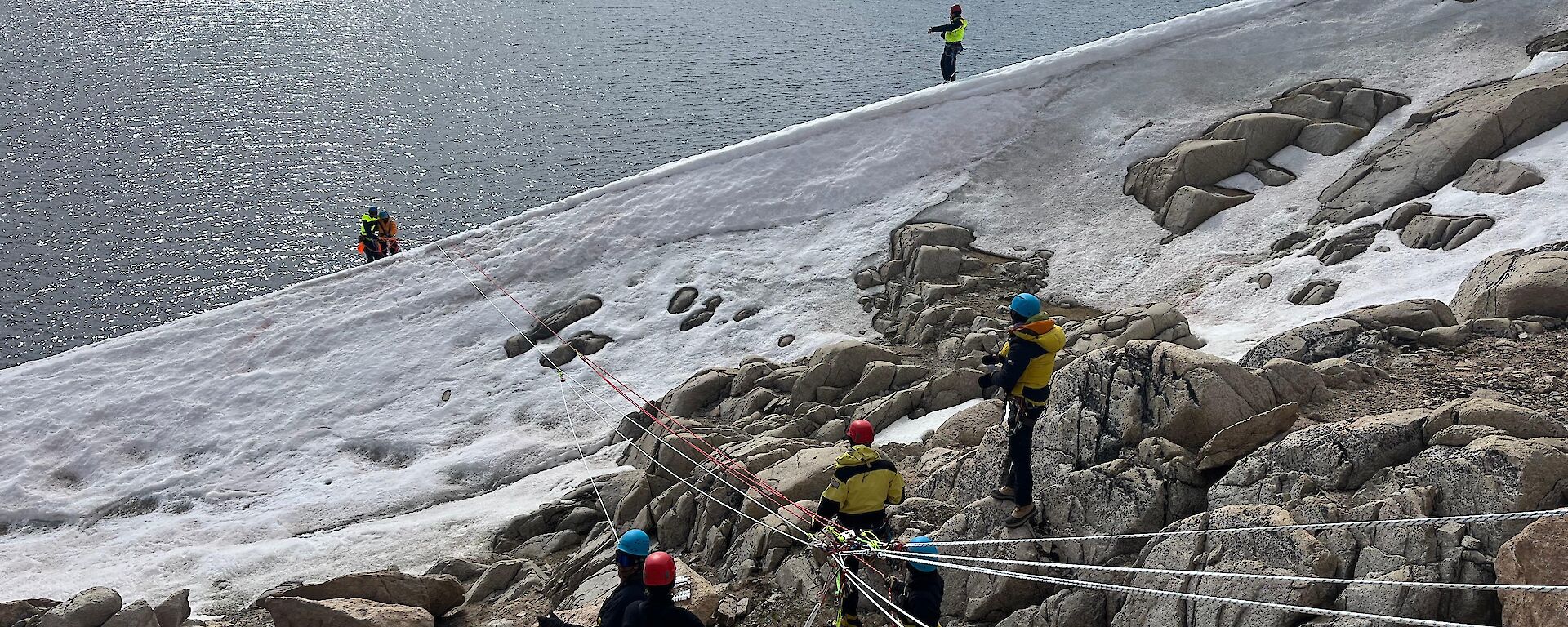 Expeditioners use ropes on the side of a rocky hill, with ice visible at the bottom of the steep hillside