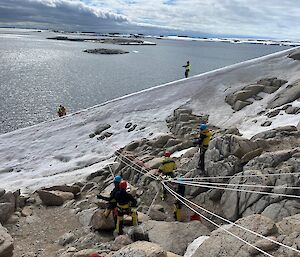 Expeditioners use ropes on the side of a rocky hill, with ice visible at the bottom of the steep hillside