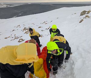 Expeditioners in yellow jackets and black pants in Antarctica carrying a patient in a red bag in Antarctica during a training drill