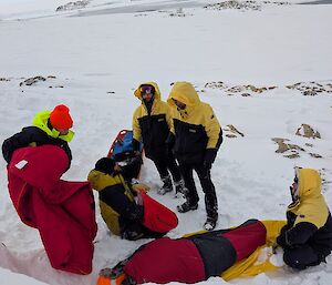 Expeditioners in yellow jackets and black pants in Antarctica treating a patient in a red bag in Antarctica during a training drill