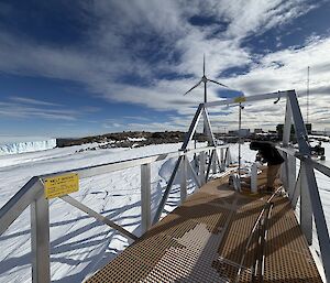 Picture of a metal bridge-like structure or pontoon, with ice, rocks and a wind turbine in the background.