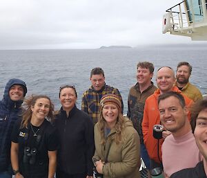 Ten people posing for a selfie on the deck of Nuyina with a clouded view of Macquarie Island in the distance.