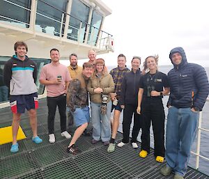 A group of people standing on the deck of RSV Nuyina in various warm and cool weather attires.
