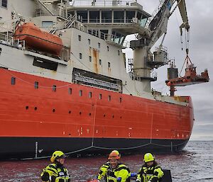 Four people in an inflateable rubber boat beside the Nuyina. Behind them a barge is being lowered from a crane off Nuyina.