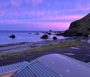 A blue and pink sunrise over the beach at Macquarie Island. The metal roof-tops of some buildings are visible in the foreground.