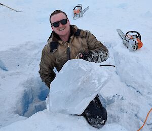 A man smiling, holding a block of ice, with a chainsaw in the background