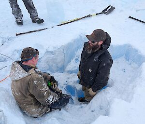 Two people sitting on the edge of a hole in the ice with shovels nearby