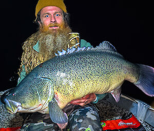A man wearing a mustard-coloured beanie is holding a very large fish towards the camera