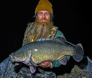 A man wearing a mustard-coloured beanie is holding a very large fish whilst sitting in a boat in the dark