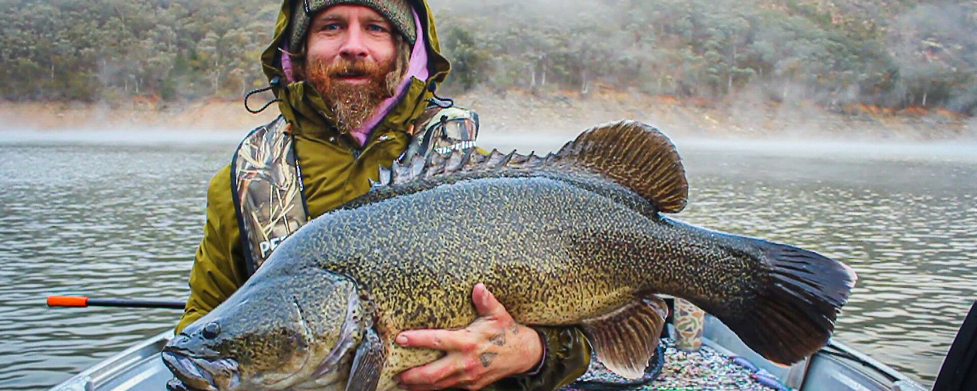 A bearded man, wearing a green jacket witht he hood up holds a very large fish