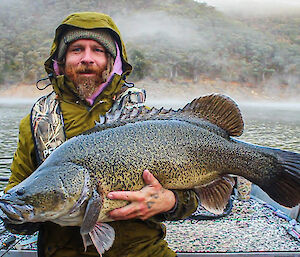 A bearded man, wearing a green jacket witht he hood up holds a very large fish