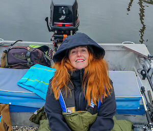 A red headed woman is wearing a navy blue jacket and a khaki green blanket whist sitting in a boat on very calm water