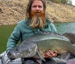 A man with dreadlocks and a red beard sits in a boat holding a very large fish across his lap