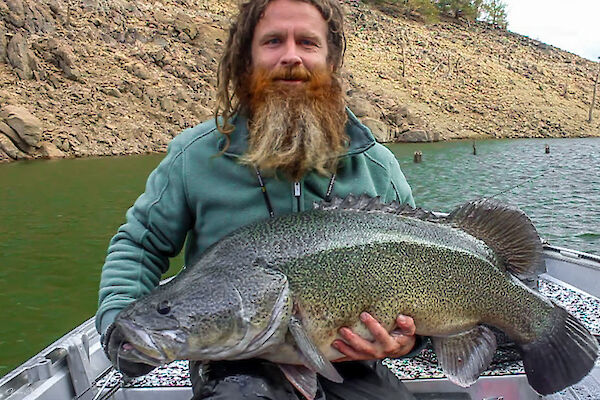 A man with dreadlocks and a red beard sits in a boat holding a very large fish across his lap