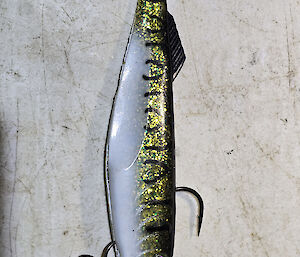 A fishing lure sits on a white bait board. It has a gold sparling back with a white underside.