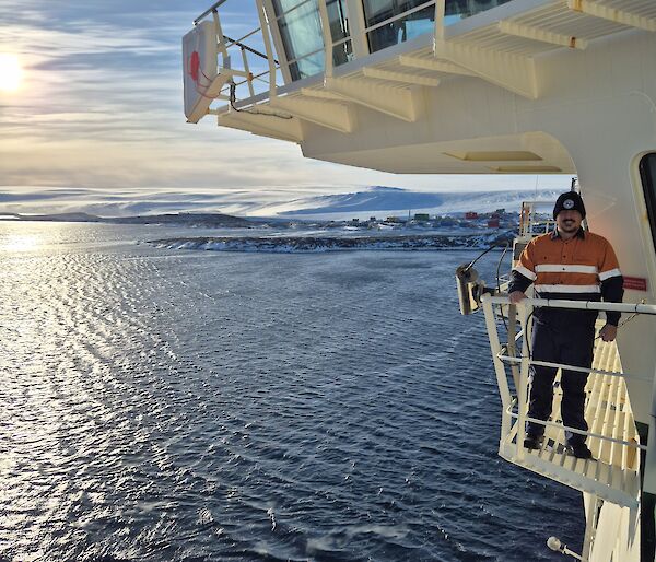 An expeditioner standing on the upper decks of the Nuyina, with Mawson Station and icy mountains in the background