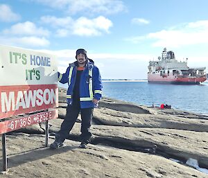 An expeditioner standing next to the "It's Home it's Mawson" sign, with the Nuyina in the background.