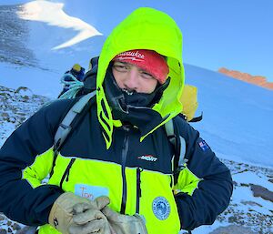 An expeditioner rugged up in field gear, standing in front of a snowy backdrop including rocks and ice cliffs.