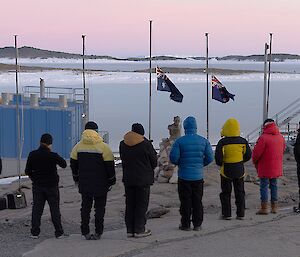 Expeditioners gathered in front of Mawson's flag poles for the ANZAC service, with sea ice in the background.
