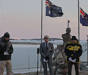 Three expeditioners in front of Mawson's flag poles delivering an address, The Ode and an ANZAC poem.