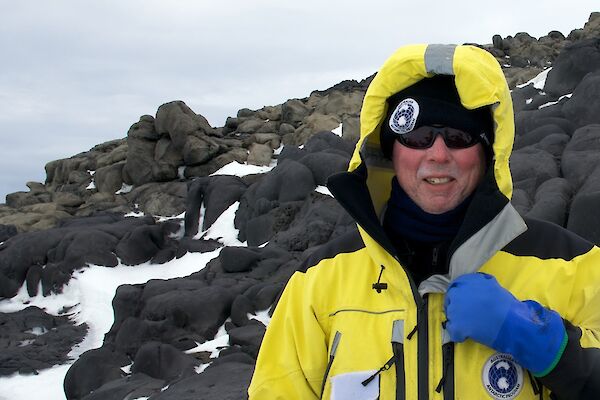 Sea ice scientist Dr Rob Massom in a yellow Antarctic jacket standing beside a rocky hill. Some snow is covering the dark rocks.