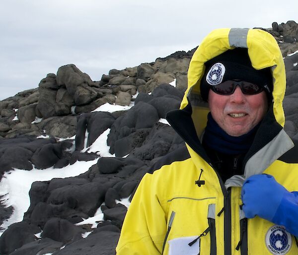 Sea ice scientist Dr Rob Massom in a yellow Antarctic jacket standing beside a rocky hill. Some snow is covering the dark rocks.