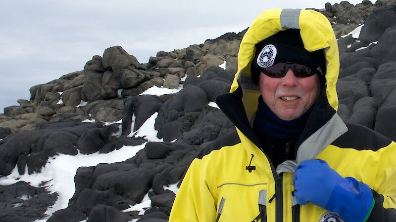 Sea ice scientist Dr Rob Massom in a yellow Antarctic jacket standing beside a rocky hill. Some snow is covering the dark rocks.