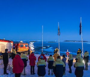 A group of expeditioners wearing large winter jackets are facing away from the camera towards a lectern, flagpoles and remembrance cross.