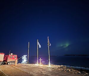 A green aurora lights a deep blue sky behind flag poles with a white cross standing between them