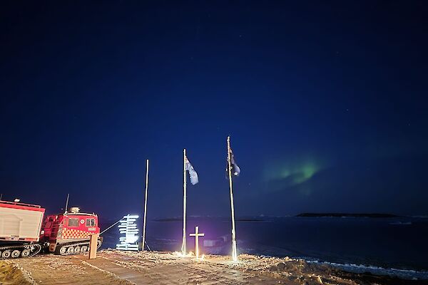 A green aurora lights a deep blue sky behind flag poles with a white cross standing between them
