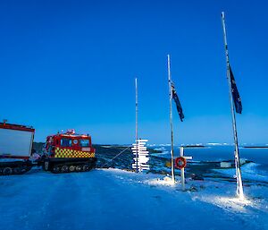 The sky is a pale blue with early dawn light. In a snowy landscape there are two flag poles, one in the foreground with the Few Zealand flag at half-mast and one with the Australian Flag at half-mast. A white cross with a poppy wreath stands between them. In the background is a red Hagglunds vehicle with emergency markings on the side.