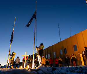 A group of expeditioners wearing winter jackets stand in front of a yellow building. Two expeditioners are raising the Australian and New Zealand flags to full mast, whilst Reveille plays.