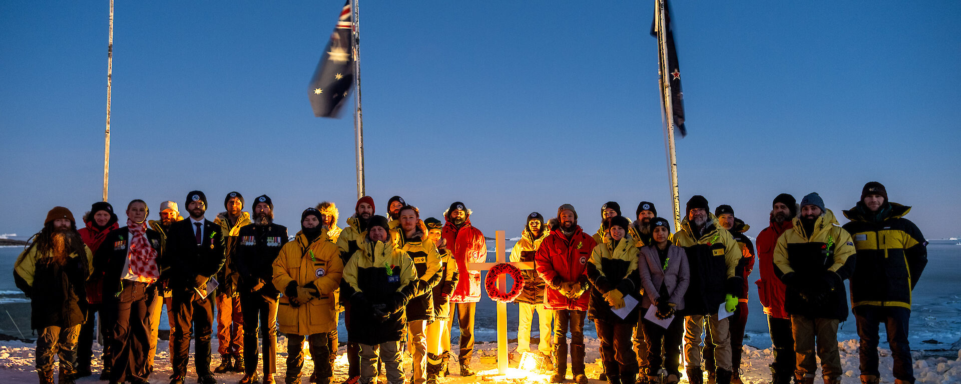 A group of expeditioners wearing heavy winter jackets stand in front of flagpoles with flags at half mast, with a white cross adorned with a poppy wreath between them