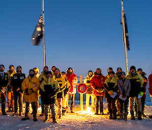A group of expeditioners wearing heavy winter jackets stand in front of flagpoles with flags at half mast, with a white cross adorned with a poppy wreath between them