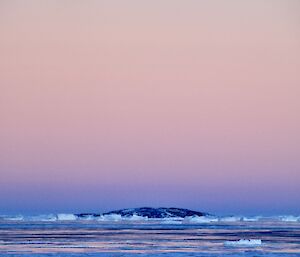 A full moon surrounded by pink and purple hues of colour, over the sea ice and water, with a rocky outcrop of an island in the distance