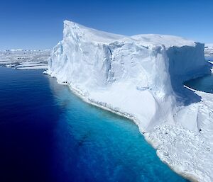 In the sky view of a large iceberg in clear-blue water, showing submerged ice below - with the feature surrounded by ice floes.